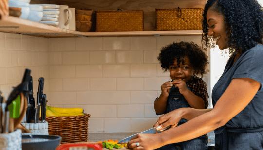 A nanny joyfully preparing a meal with a smiling child in a cozy kitchen.