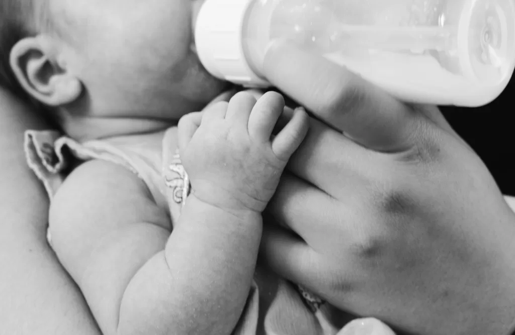 Black and white image of an infant's hand grasping an adult's finger while being bottle-fed, symbolizing care and feeding support.